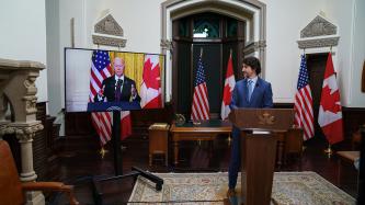 PM Trudeau stands at a podium looking at a screen of President Biden at a podium