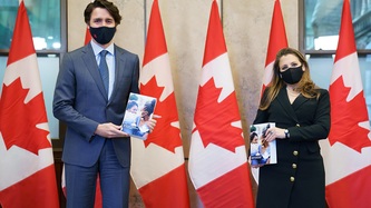 PM Trudeau and DPM Freeland stand holding a book, a row of Canadian flags behind them