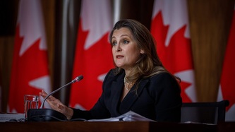 Deputy Prime Minister Chrystia Freeland sits at a microphone, a row of Canadian flags behind her