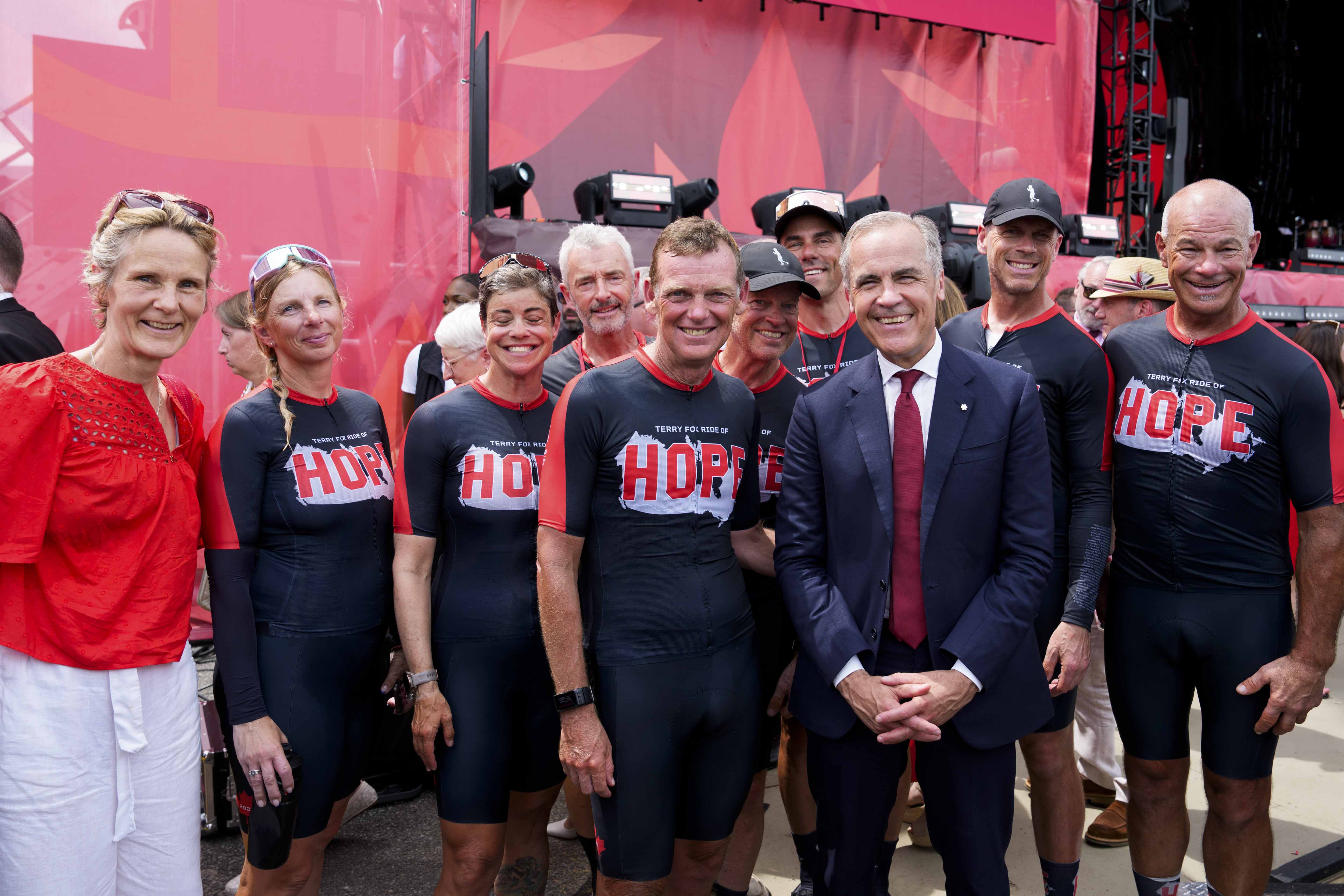 Le premier ministre Carney et Diana Fox Carney sont debout accompagnés de Darrell Fox et d’autres cyclistes à la cérémonie de la fête du Canada.