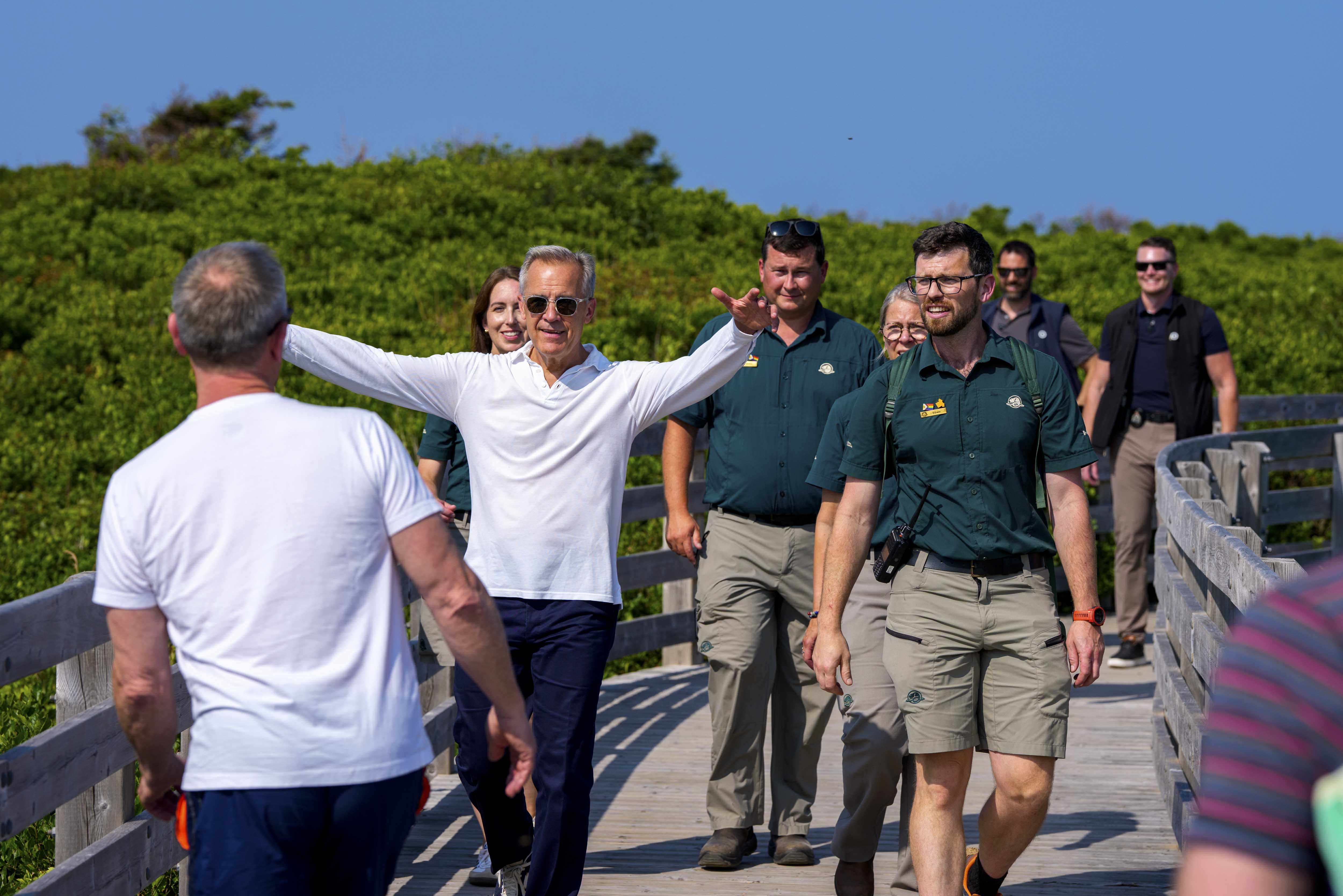 Prime Minister Mark Carney, accompanied by Parks Canada employees, walks toward a man with arms opened at PEI National Park.