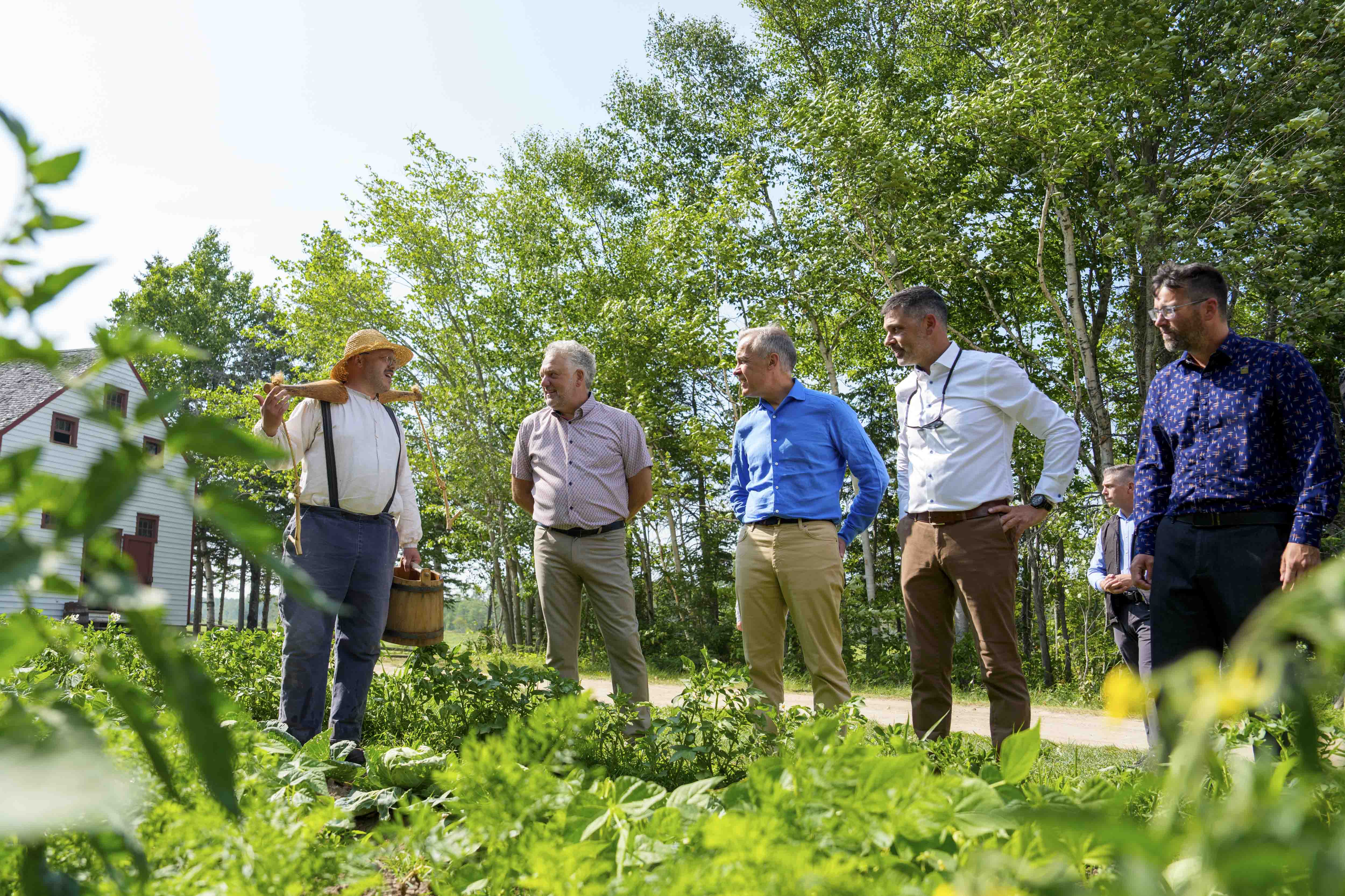 Le premier ministre Mark Carney et d’autres personnes sont debout et discutent avec un agriculteur dans un jardin au Village historique acadien.