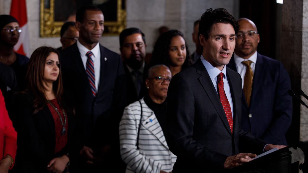 Inside Parliament, community leaders and members of parliament stand behind Prime Minister Trudeau while he makes an announcement