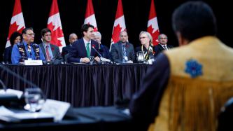 Prime Minister Justin Trudeau sits in front of a microphone and delivers remarks while Minister Carolyn Bennett and other officials and ministers look on
