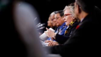 Minister Carolyn Bennett sits in front of a microphone and delivers remarks while officials and ministers look on