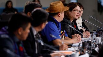 Minister Jane Philpott sits in front of a microphone and delivers remarks while officials and ministers look on