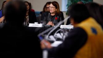 Minister Jane Philpott sits in front of a microphone and delivers remarks while officials and ministers look on
