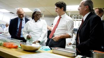 Prime Minister Justin Trudeau laughs with a chef at Cuisine-Atout in a kitchen while MP David Lametti and kitchen staff look on