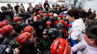 Prime Minister Justin Trudeau leans in from outside the rink while speaking with a crowd of youth on the ice gathered near him