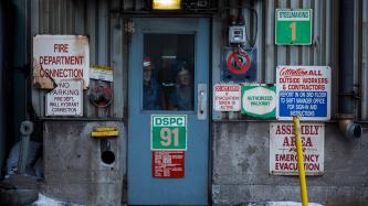 Algoma steel workers in hard hats peer out from the glass window of an entrance door surrounded by several warning and instruction signs