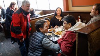 Prime Minister Justin Trudeau crouches next to a table to talk with customers of Muio's Restaurant while MP Terry Sheehan stands at his side.