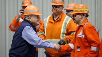 Minister Ralph Goodale wears a hard hat and safety goggles and shakes hands with an EVRAZ Regina employee alongside a few others