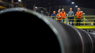 Prime Minister Justin Trudeau, Minister Ralph Goodale and three uniformed employees wear hard hats and safety goggles as they look out at the EVRAZ Regina facility from a footbridge above