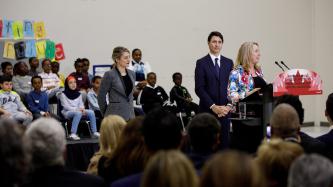 A woman addresses the audience at a school from a podium with a sign that reads “Strengthening our official languages” in both official languages while Prime Minister Justin Trudeau and Minister Mélanie Joly stand behind her.