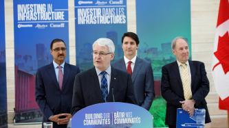 Minister Marc Garneau speaks at the podium alongside PM Trudeau and Minister Amarjeet Sohi