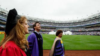 Prime Minister Justin Trudeau looks up at the crowd at Yankee Stadium flanked by two women, all in doctoral regalia