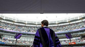 Prime Minister Justin Trudeau stands at a podium in doctoral regalia and looks at the crowd in Yankee Stadium