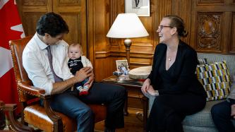 Prime Minister Justin Trudeau sits across from Minister Karina Gould holding her baby in the Prime Minister’s Office