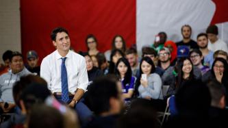 PM Trudeau sits in a room surrounded by a crowd