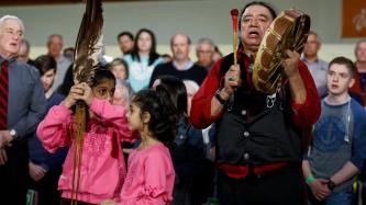 An indigenous man bangs a drum while two girls hold a feather
