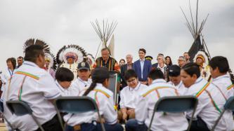 A man speaks at a podium in front of PM Trudeau and Ministers Bennett and Goodale