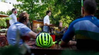 People sit at tables under trees with PM Trudeau at a podium, MP Amos looks on