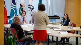 A woman stands at a table where PM Trudeau and two women look on