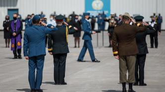 Military officers stand in salute position as a colleague bears a service hat on a pillow