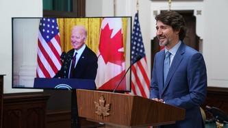 Le PM Trudeau sourit derrière un lutrin; à l’écran, le président Biden sourit derrière un lutrin
