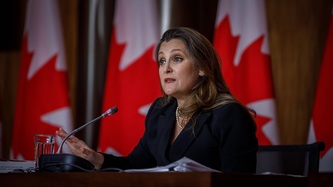 La vice-première ministre Freeland est assise derrière un micro devant des drapeaux canadiens