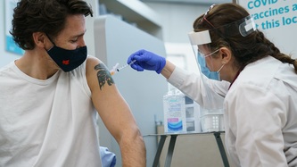 Prime Minister Trudeau looks on as a woman in a white cloak administers a needle to his arm