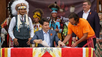 PM Trudeau holds papers at a table, Cowessess First Nation Chief Cadmus Delorme and others look on