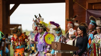 A woman stands at a podium with a large group of Cowessess First Nation people surrounding her