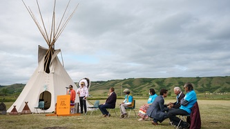 Prime Minister Justin Trudeau kneels and holds hands with someone sitting in a chair in a field