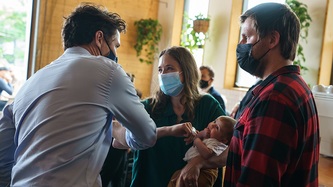 PM Trudeau and a woman holding a baby exchange an elbow bump as a man looks on