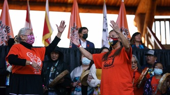 Two women wearing orange t-shirts motion with their hands toward the sky in front of hand drummers