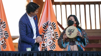 PM Trudeau stands looking at Chief Casimir holding a painted hummingbird hand drum