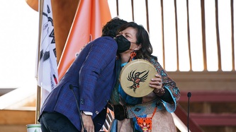 PM Trudeau and Chief Casimir hug as she holds a painted hummingbird hand drum 