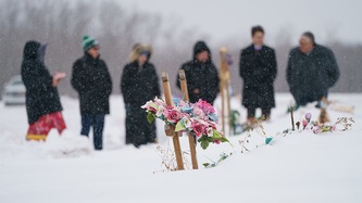 Des couronnes de fleurs reposent sur des croix de bois. À l'arrière-plan, des gens sont debout.