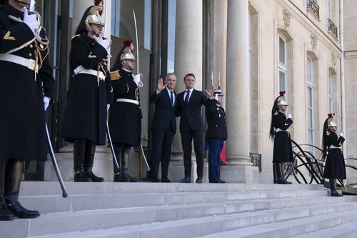 Prime Minister Carney and President Macron wave to media outside the Élysée Palace. Guards are standing on both sides.