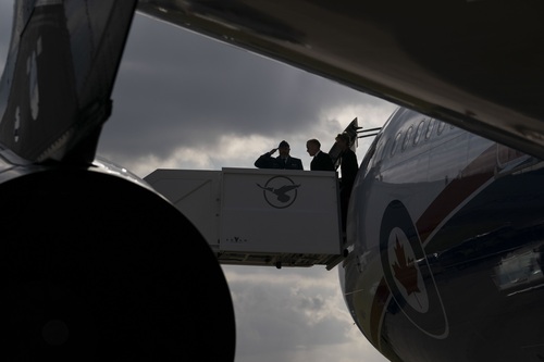 The silhouettes of Prime Minister Carney disembarking an aircraft in London and a man standing at attention and saluting.