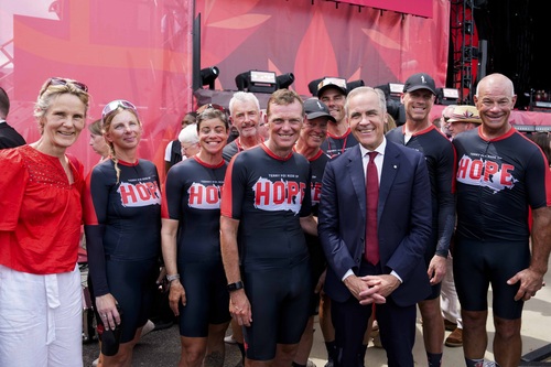 Prime Minister Carney and Diana Fox Carney standing with Darrell Fox and other cyclists at the national Canada Day ceremony.