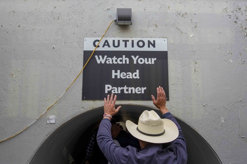 Prime Minister Carney raises his arms below a sign that reads “Caution: Watch Your Head Partner” at the Calgary Stampede.