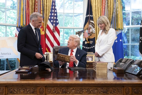 Prime Minister Carney and Minister Joly stand beside President Trump who is sitting at the Resolute Desk. 