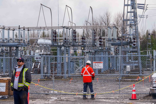 Two construction workers in safety gear stand near a chain link fence on the grounds of the BC Hydro Skeena Substation, wi...