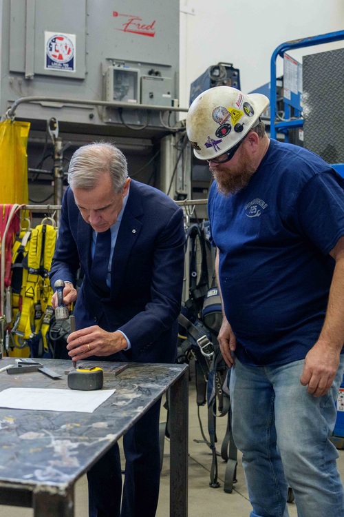 Le premier ministre Mark Carney grave des marques sur un morceau de métal à l'aide d'un marteau, tandis qu'un travailleur ...