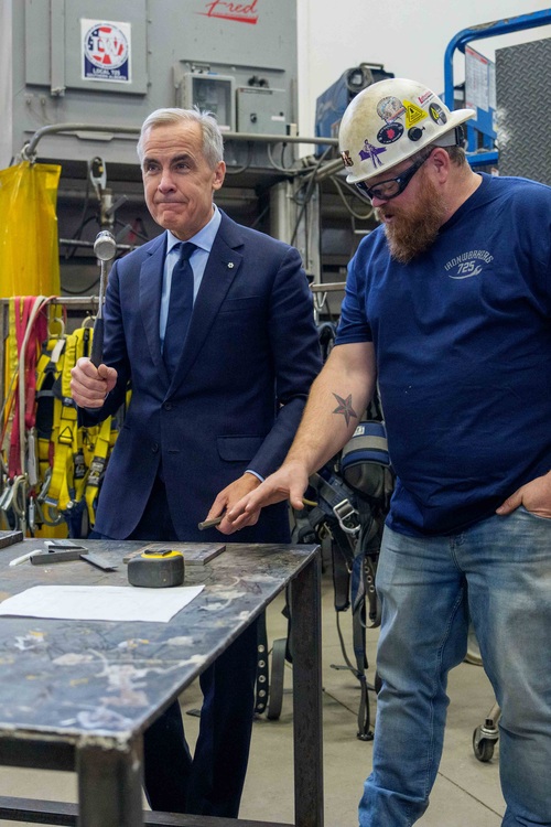 Le premier ministre Mark Carney se tient debout devant un établi en métal en tenant un marteau, tandis qu'un travailleur s...