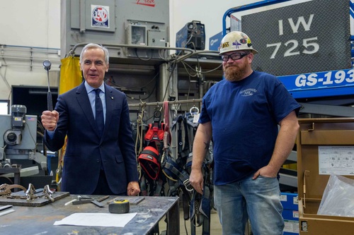 Le premier ministre Mark Carney se tient debout devant un établi en métal en tenant un marteau, tandis qu'un travailleur s...